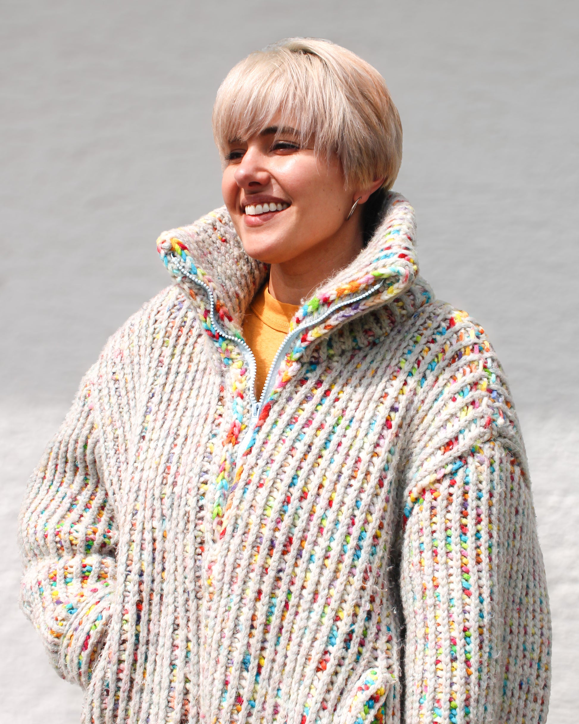 Pattern Designer smiling while wearing a size MEDIUM Brioche Crochet Pullover in the fruity-pebbles colorway. Statement collar is shown half zipped layered over an orange shirt for a layered look in a joyful colorway. 