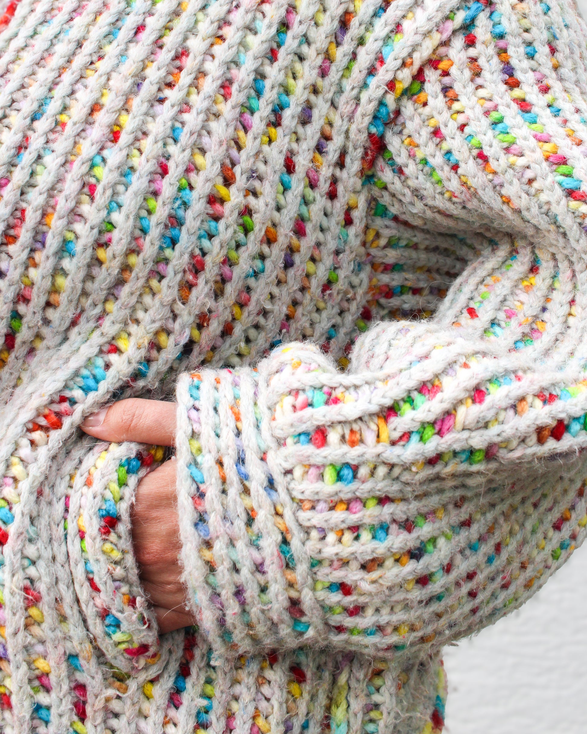 Close-up detail of the chunky two-color brioche stitch texture in the fruity-pebbles colorway. Shot shows the perpendicular cuff and a hand in the 'faux welt' entry to the deep pocket. Rainbow speckled yarn through a light gray-blue base. 