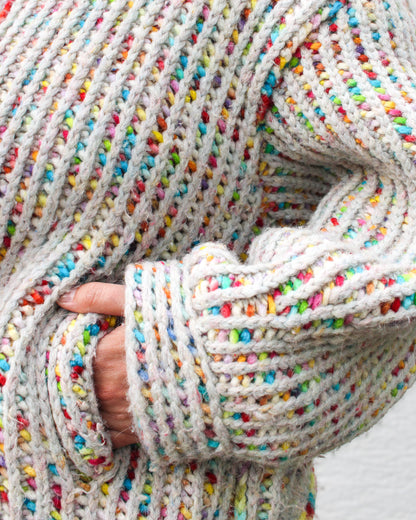 Close-up detail of the chunky two-color brioche stitch texture in the fruity-pebbles colorway. Shot shows the perpendicular cuff and a hand in the 'faux welt' entry to the deep pocket. Rainbow speckled yarn through a light gray-blue base. 